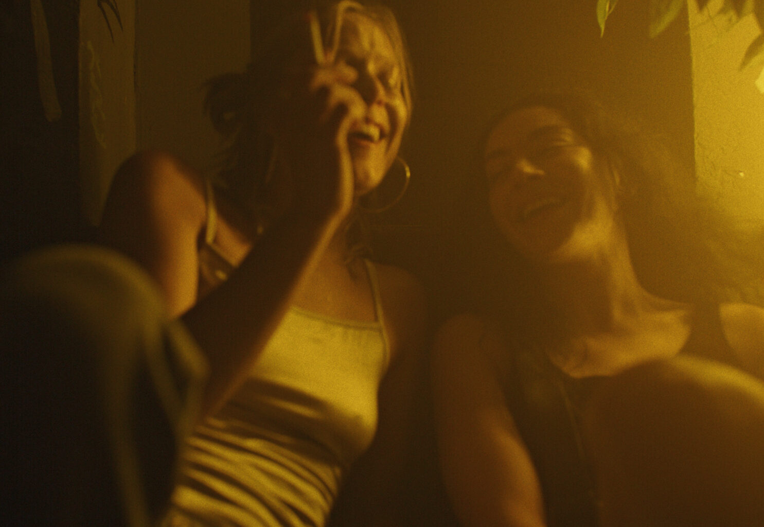 Still from the Erupcja showing two women in casual clothes laughing together as one smokes a cigarette. The film still is bathed in yellow light