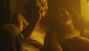 Still from the Erupcja showing two women in casual clothes laughing together as one smokes a cigarette. The film still is bathed in yellow light