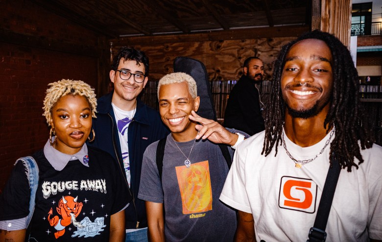 Tampa Bay songwriter Fessi K (Zoe Robinson) and her band members smile together in a candid indoor portrait. Fessi K wears a black "Gouge Away" graphic t-shirt over a collared shirt, and the group is dressed in casual streetwear, reflecting their presence in the local DIY and punk scenes.
