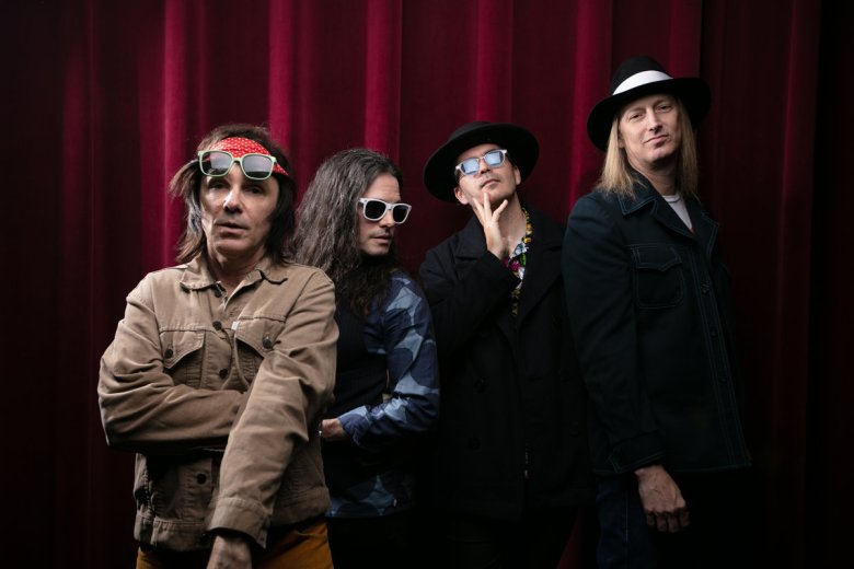 A stylized group portrait of the four-member rock band Florida. On the far left, frontman Billy Summer stands with arms crossed, wearing a tan trucker jacket, a red patterned headband, and large aviator sunglasses with a red-and-white frame. To their right, three other band members pose against a deep red velvet stage curtain. The second member has long dark hair and dark glasses; the third wears a black hat and coat with a floral shirt; and the fourth, on the far right, wears a white fedora-style hat and a dark jacket. The lighting is dramatic, casting a moody glow over the group.