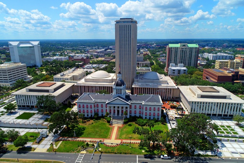 An aerial view of the Florida State Capitol complex in Tallahassee. The historic 1845 Capitol building with its grey dome and candy-striped awnings sits in the foreground, directly in front of the modern 22-story Executive Office Building tower. The surrounding plaza, legislative wings, and downtown Tallahassee skyline are visible under a blue sky with scattered clouds.
