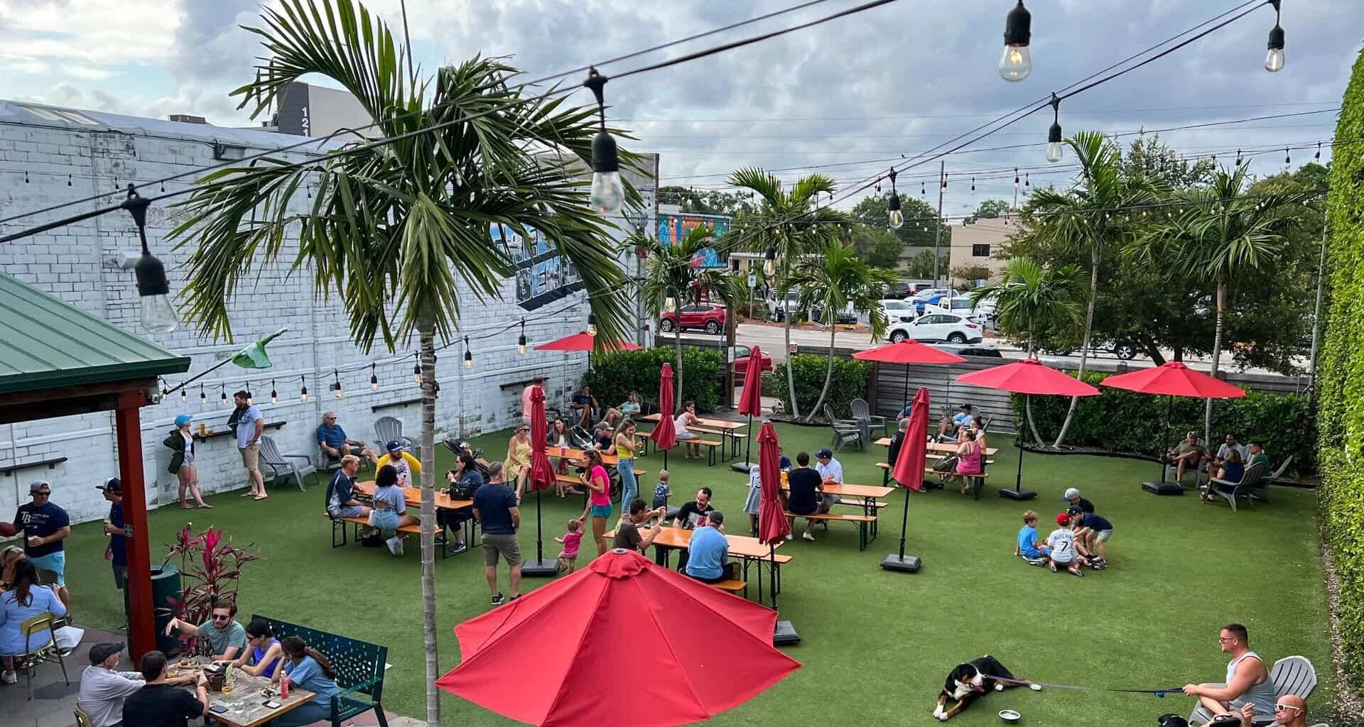 Outdoor photograph of a beer garden with people sitting at tables under red umbrellas on artificial grass, surrounded by palm trees and string lights overhead.
