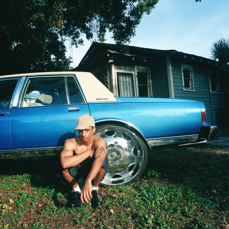 Musician Gabriel Jacoby crouched in front of a blue vintage car with large chrome rims. The person is shirtless, wearing a tan cap and black shoes, posed in front of a blue wooden house with palm trees under a clear blue sky.