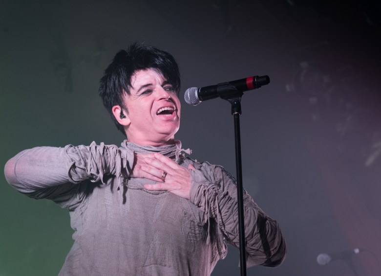 Gary Numan performing live on stage, singing into a microphone with one hand over the chest. The artist has short black hair and dark eye makeup, wearing a distressed, grey long-sleeve tunic with shredded fabric details against a dark, smoky background.