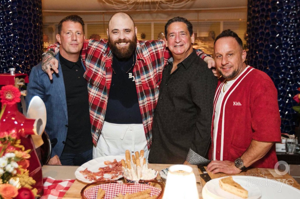 photo of four men posing together at a party with bread and prosciutto on the table in front of them