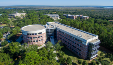 Aerial shot of UWF's Hal Marcus College of Science and Engineering building in the shape of a zero and one, surrounded by greenery and a blue sky.