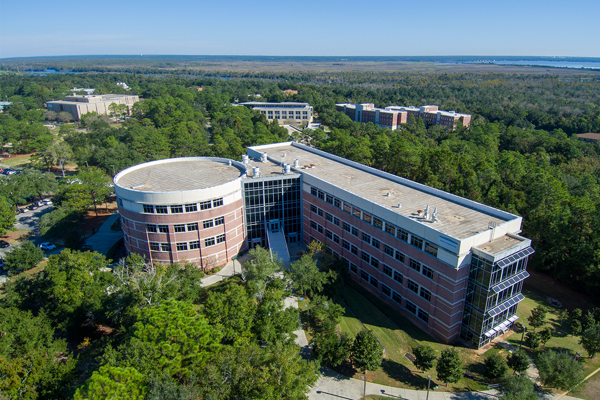 Aerial shot of UWF's Hal Marcus College of Science and Engineering building in the shape of a zero and one, surrounded by greenery and a blue sky.