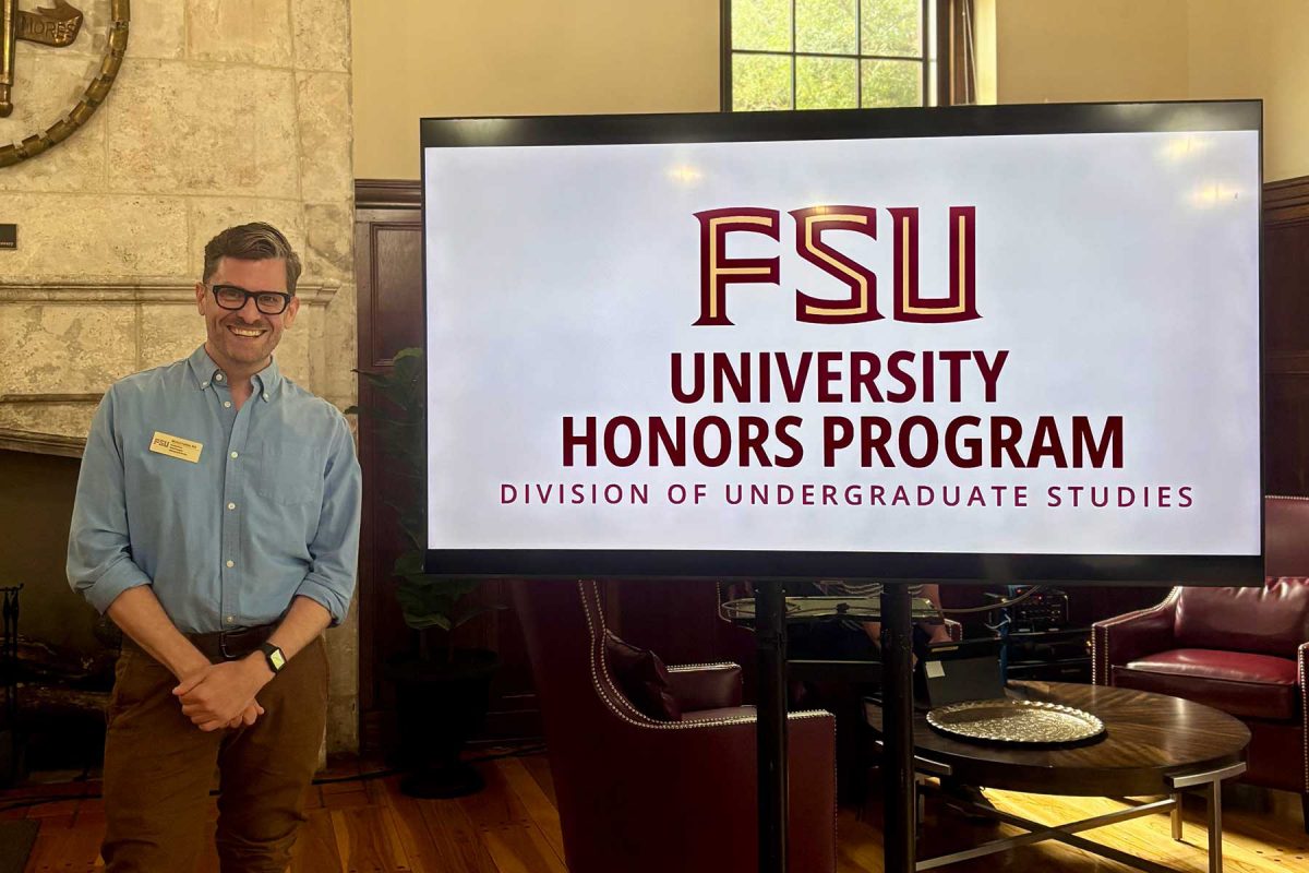 Michael David Franklin, Associate Director of Honors in the Major, smiles next to a digital display for the FSU University Honors Program in the Beth Moor Lounge.