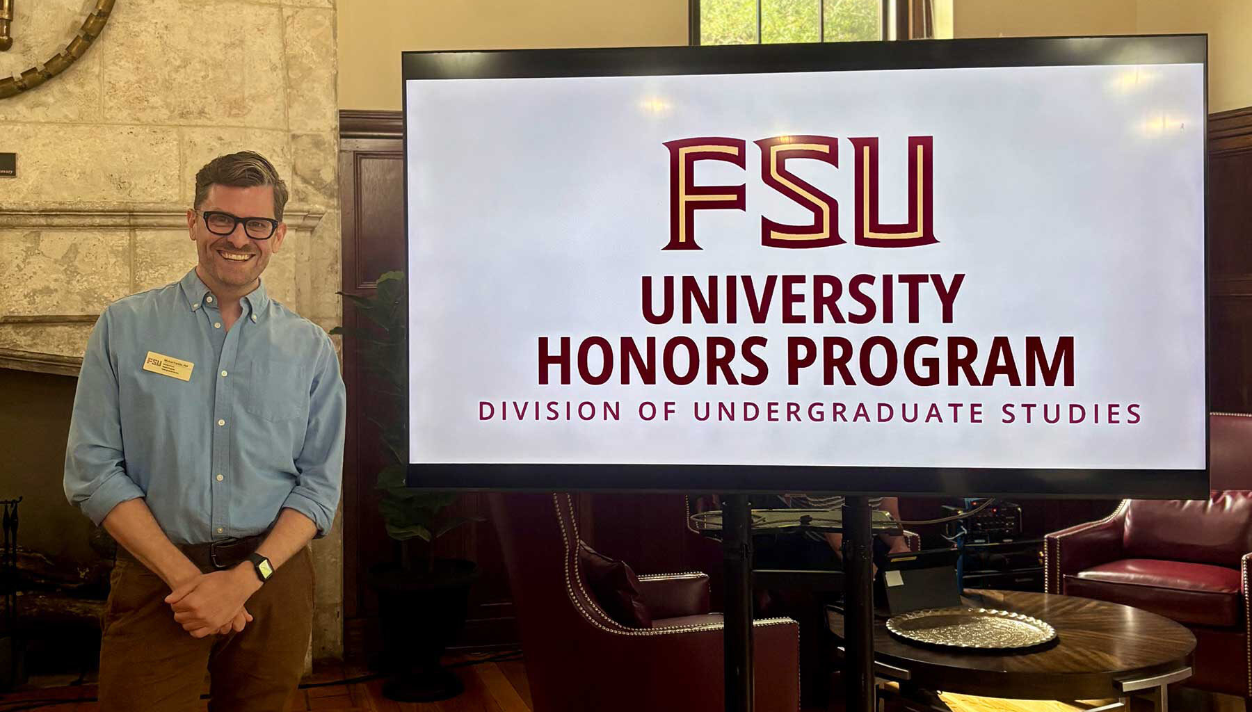 Michael David Franklin, Associate Director of Honors in the Major, smiles next to a digital display for the FSU University Honors Program in the Beth Moor Lounge.