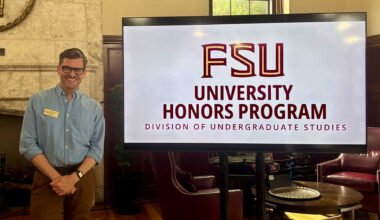 Michael David Franklin, Associate Director of Honors in the Major, smiles next to a digital display for the FSU University Honors Program in the Beth Moor Lounge.