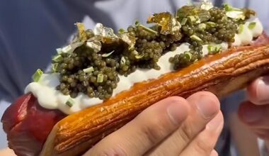 A man holding a Wagyu hot dog in a croissant bun topped with caviar, creme fraiche, and gold flakes