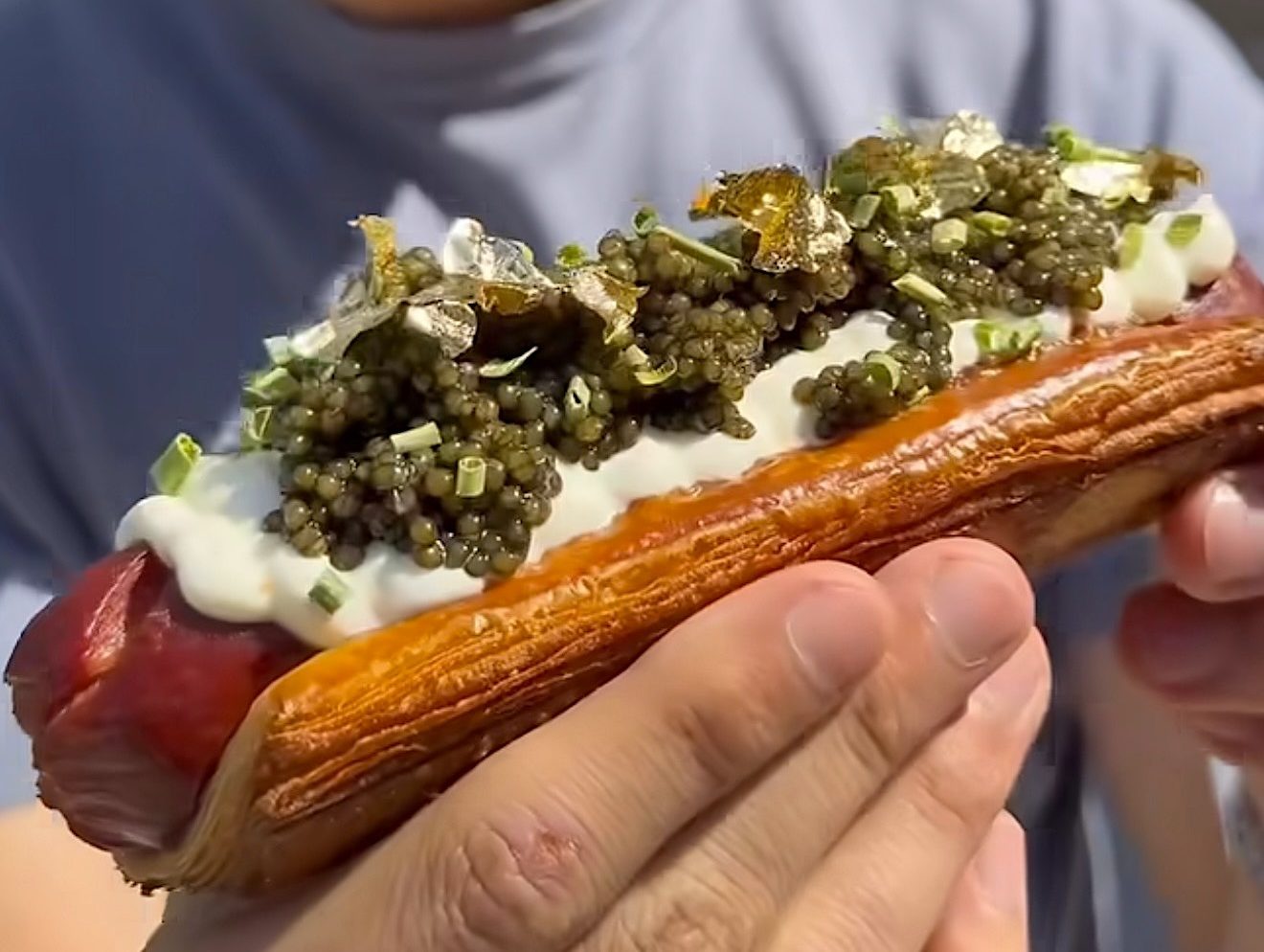 A man holding a Wagyu hot dog in a croissant bun topped with caviar, creme fraiche, and gold flakes