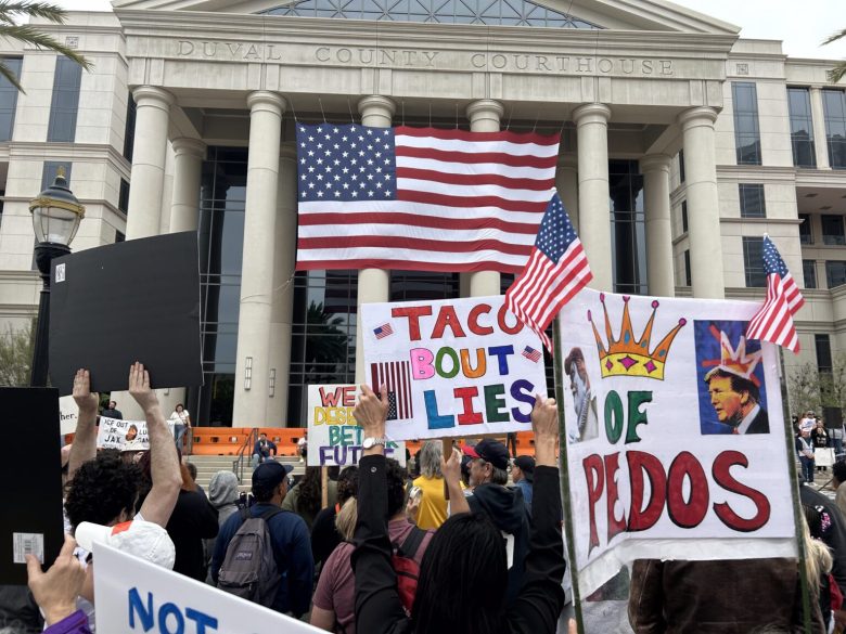 Protesters gather outside the Duval County Courthouse in Jacksonville, Florida, holding signs including one that reads "Taco 'Bout Lies" next to a large American flag draped between courthouse columns.