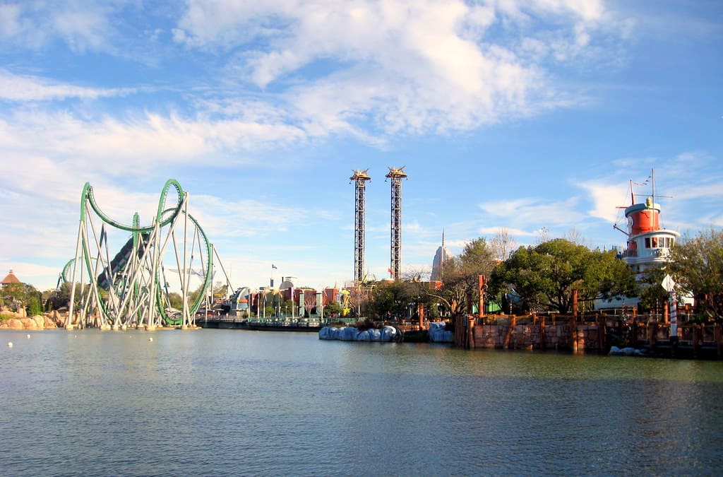 A theme park scene with a large green roller coaster, two tall drop towers, and the Seuss Landing Café near a red and white lighthouse beside a lake under a partly cloudy blue sky. Trees and buildings line the water’s edge.