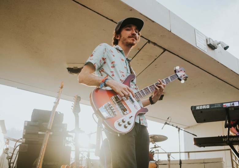 Musician Jaron Jammer playing a multicolored electric guitar on an outdoor stage at a low angle. The person wears a light blue flamingo-print shirt and a dark baseball cap, with stage monitors and a Korg keyboard visible in the background under bright, hazy sunlight.