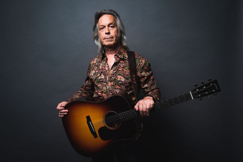 A studio portrait of country and Americana singer-songwriter Jim Lauderdale against a dark grey background. The person has silver, shoulder-length hair and wears a vibrant, multi-colored floral western-style shirt with pearl snap buttons. They are holding a sunburst-finish acoustic guitar with a dark leather strap, looking directly at the camera with a steady, neutral expression.