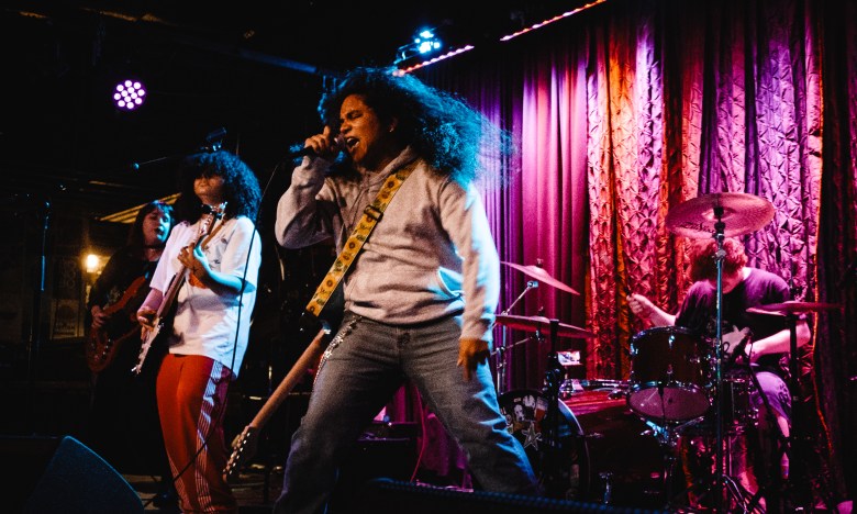 Tampa-based punk band Just Courtesy performing live on stage. The lead singer, with voluminous curly hair and a grey hoodie, sings passionately into a microphone while a guitarist in red track pants and a drummer perform under dramatic purple and red stage lighting.