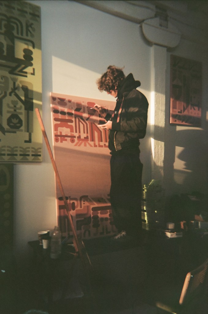 A candid photograph of artist Keya Tama in a sunlit studio. The artist, with dark curly hair and wearing a dark puffer jacket, stands in profile looking down at his phone. Warm, late-afternoon sunlight streams across the room, illuminating large-scale paintings on the wall that feature his signature red, black, and cream graphic style. The scene is framed by a heavy black border, creating a cinematic, intimate feel.