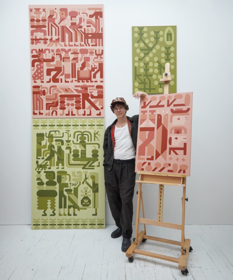 Artist Keya Tama stands in a white studio between three of his paintings. He wears a black jacket over a white tank top, black trousers, and a patterned bucket hat. To his left, two large vertical canvases are stacked; the top one features red and pink graphic figures, while the bottom shows similar figures in green and olive. To his right, a smaller pink and red painting sits on a wooden easel. The artist rests his hand on the easel, looking toward the camera against a stark white floor and walls.