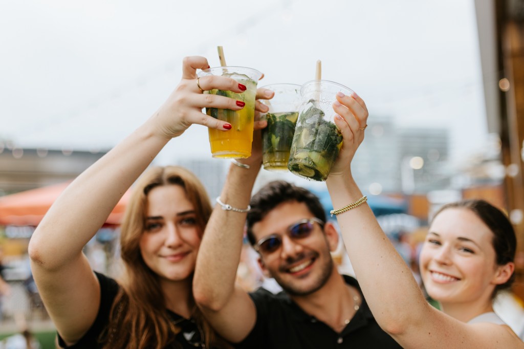 three people posing for the camera outside and smiling while cheersing with three drinks in plastic cups