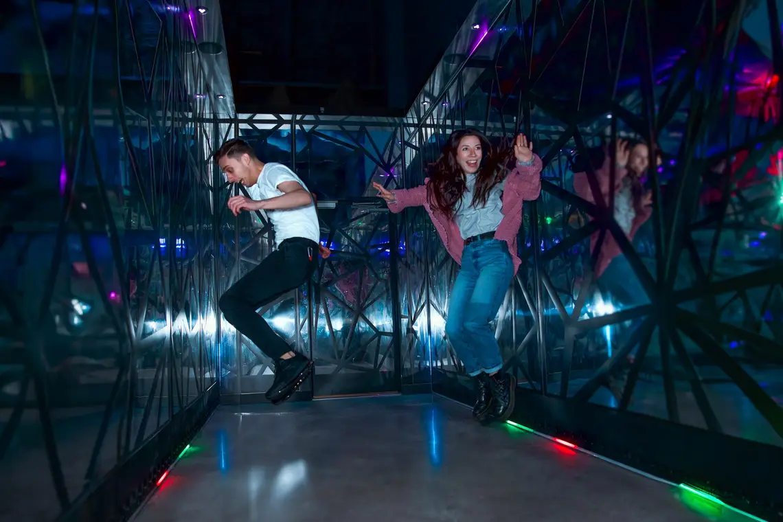 Two people joyfully jump and smile in a vibrant, mirrored room with neon lights—like a futuristic Disney attraction interior.