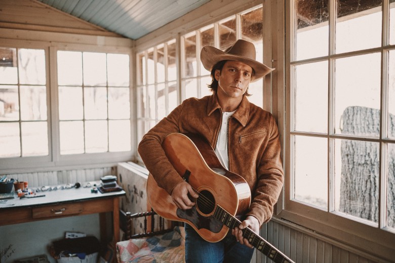Musician Lukas Nelson wearing a brown suede jacket, a white t-shirt, and a tan cowboy hat. The person is holding an acoustic guitar and leaning against a window frame inside a rustic, sunlit wooden room with multiple windows.