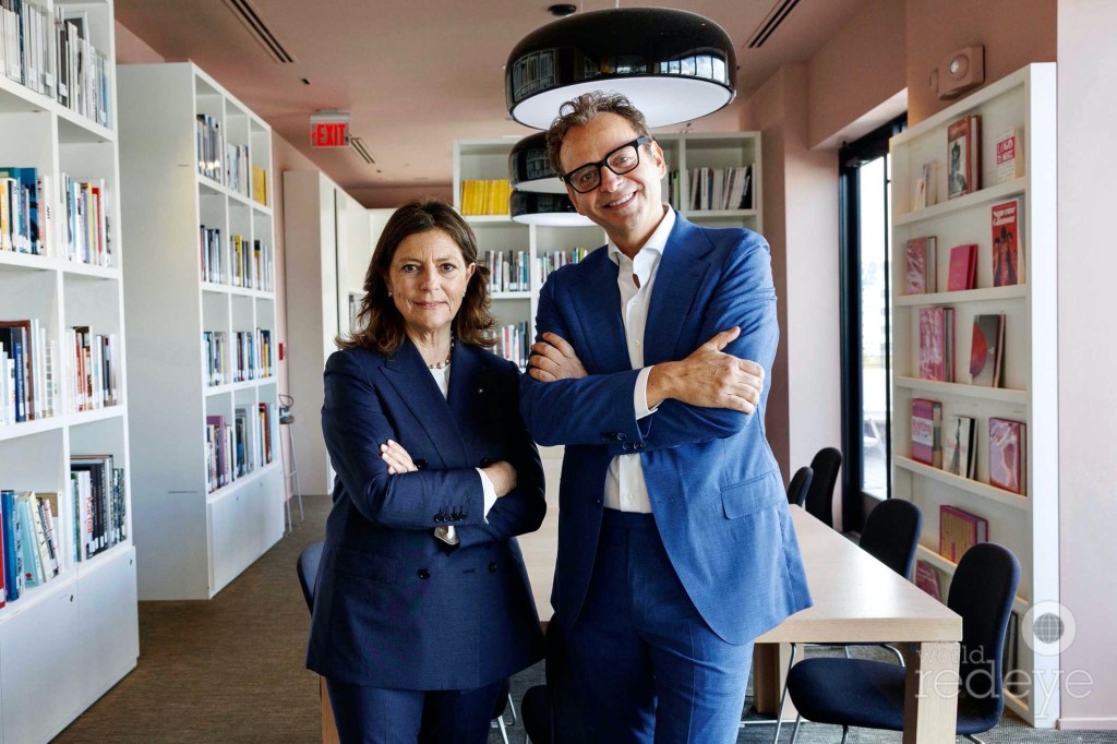 photo of a woman and man in matching blue suits posing with their arms crossed inside a studio with white bookshelves and a conference table behind them