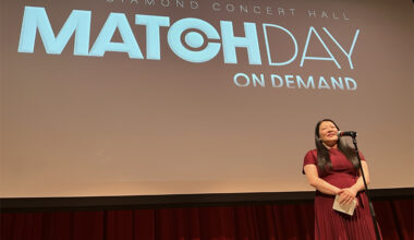 A woman in a red dress stands in front of a microphone. A screen behind her reads