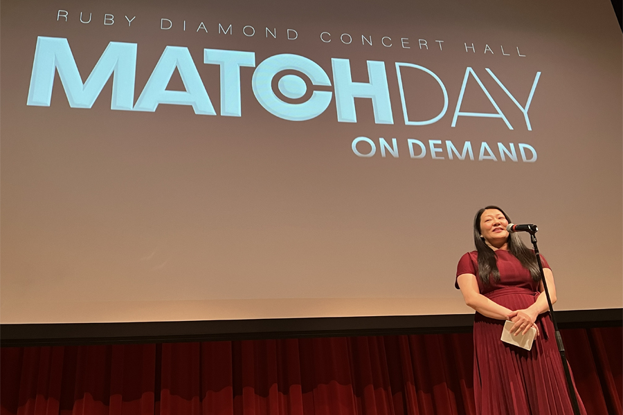 A woman in a red dress stands in front of a microphone. A screen behind her reads