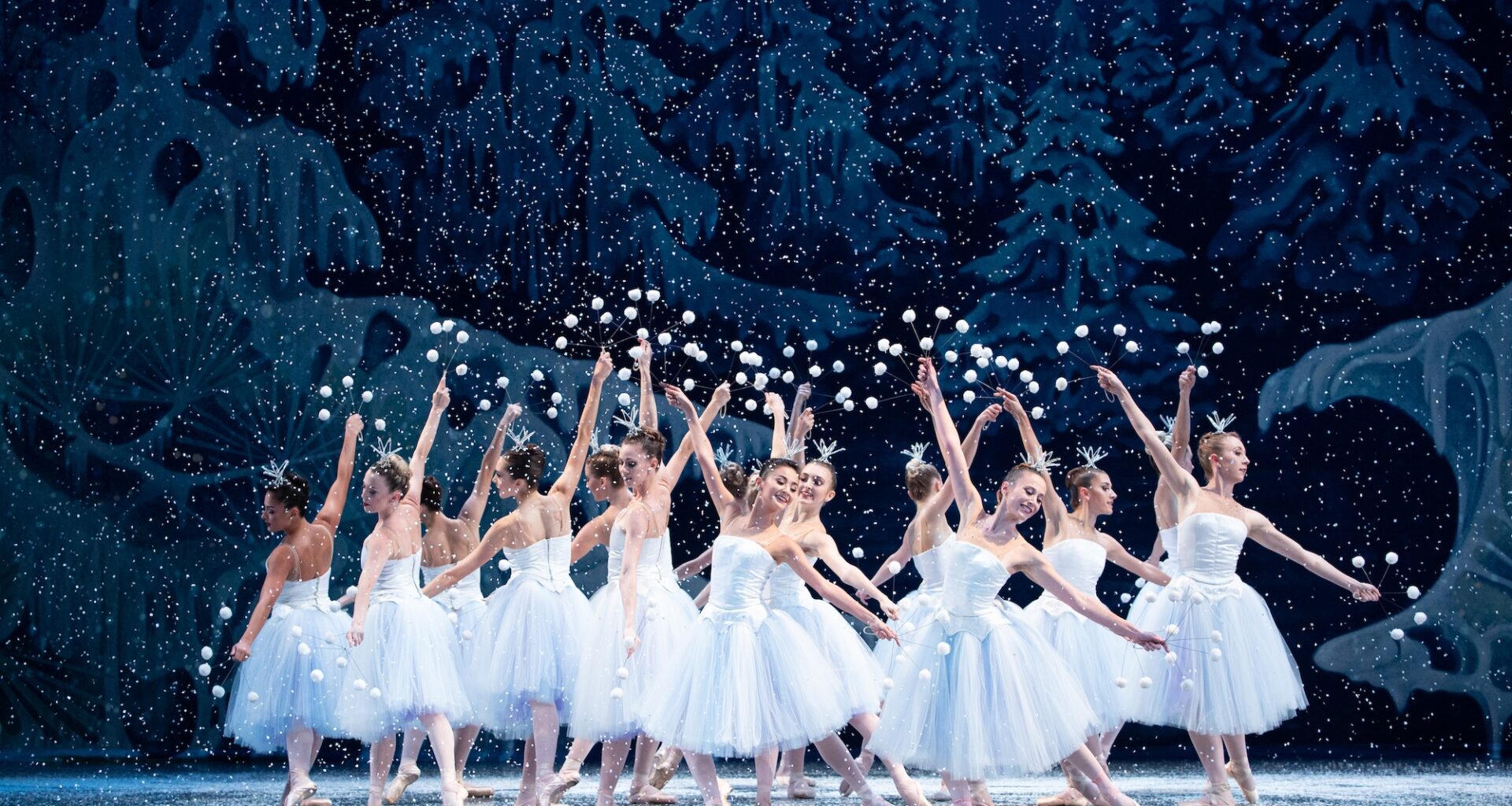 Photo of about a dozen ballerinas dancing on a stage in tiaras, tulle skirts, and pointe shoes. Fake snow appears to be falling all around them