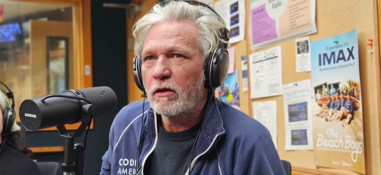 Michael Bishop, founder of the Tampa Monitor, speaking into a studio microphone at WMNF 88.5 FM. The person has grey hair and a beard, wearing headphones and a dark blue jacket. The background features a corkboard with various posters, including one for The Beach Boys and an IMAX film.
