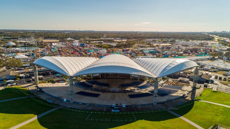 High-angle aerial view of the MIDFLORIDA Credit Union Amphitheatre in Tampa. The iconic white tensile fabric roof covers the stage and seated pavilion, surrounded by green lawns and the sprawling parking areas of the Florida State Fairgrounds under a clear blue sky.