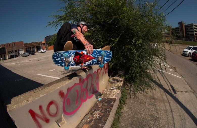 Professional skateboarder and musician Mike Vallely performing a trick on a concrete barrier with graffiti. The person is mid-air, crouching over a blue patterned skateboard; the background shows a gravel path, green bushes, and a clear sky.