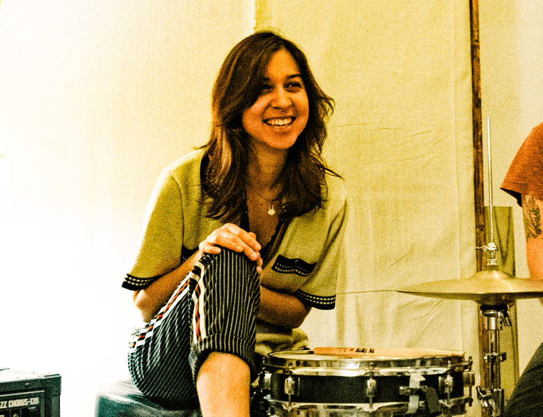 Natalie Depergola, a musician with long dark hair, smiling while sitting behind a drum kit in a studio setting next to a Roland Jazz Chorus amplifier.
