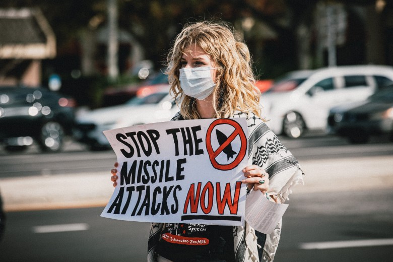 A protester in Tampa stands on a street corner, holding a handmade sign that reads "STOP THE MISSILE ATTACKS NOW." She is wearing a white medical mask and a patterned keffiyeh-style scarf, with out-of-focus cars and street background.