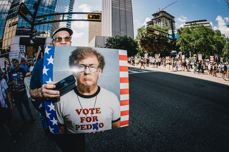 A protester in Downtown Tampa holds a sign featuring a stylized image of a man in a "Vote for Pedro" shirt, edited to say "Vote for Ped*o," during a "No Kings" rally on March 28, 2026. Tall city buildings and a crowd of demonstrators are visible in the background under a clear blue sky.