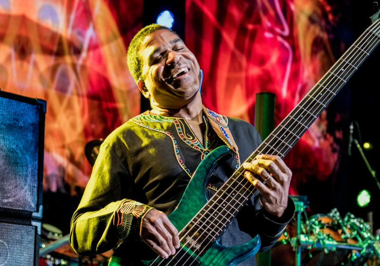 A vibrant, live performance shot of a musician playing a teal-colored six-string electric bass guitar. The person is smiling broadly with their eyes closed, wearing a black long-sleeved shirt with an intricately embroidered, colorful "dashiki-style" collar. They are performing on a stage with a bright, abstract red and yellow light display in the background, with a stack of black amplifiers visible on the left.