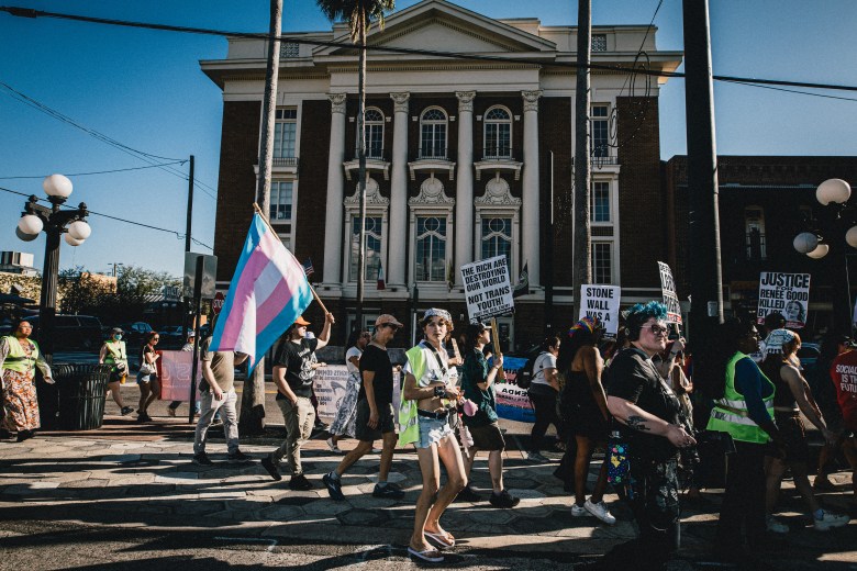 "A group of people marching during the People's Pride Coalition event in Ybor City. A person in the foreground carries a large blue, pink, and white Transgender Pride flag. The marchers are positioned in front of the historic, white-pillared Italian Club building under a clear, sunny sky.