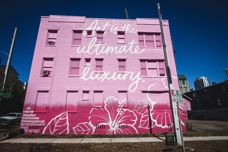 A pink-painted vintage apartment building in downtown St. Petersburg features a large white mural reading 'Art is the ultimate luxury' with hibiscus flowers, created as a temporary installation before the 1920s-era affordable housing is demolished for the 29-story Roche Bobois luxury condo tower.