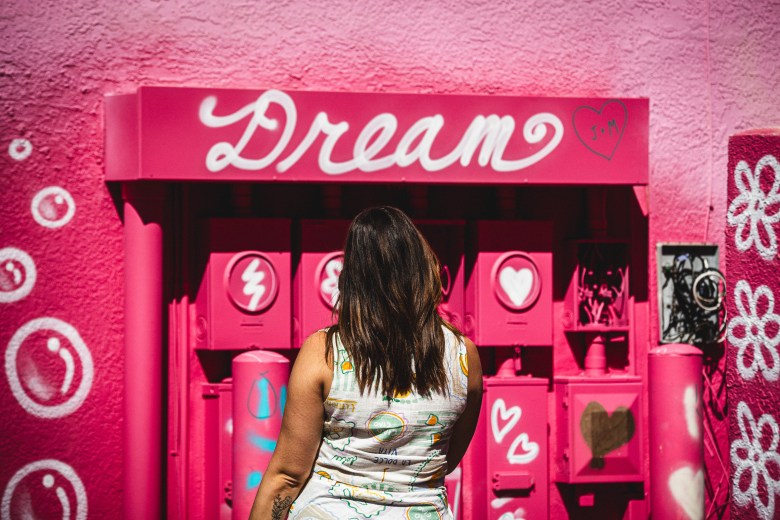 A woman with her back to the camera looks at a pink-painted apartment facade in St. Petersburg, featuring a 'Dream' sign above the doorway, pink-painted mailboxes with white heart and lightning bolt icons, and painted bubbles.