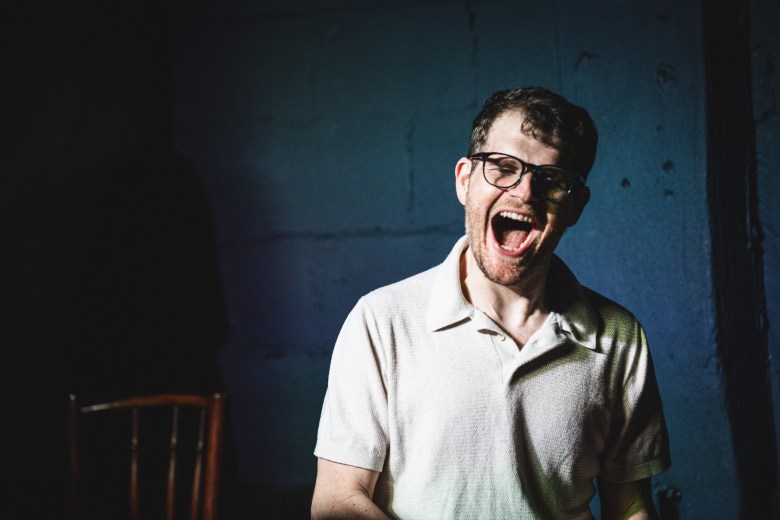 "A high-contrast portrait of Eddie Geller against a dark blue textured wall. They have short dark hair and wear black-rimmed glasses and a light-colored polo shirt. They are captured in a candid, high-energy moment with their mouth wide open as if shouting or laughing loudly. A dark wooden chair is partially visible in the shadows to the left.