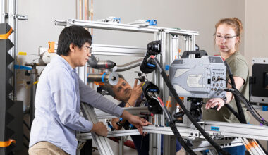 Three researchers work together on a complex experimental rig, adjusting sensors and structural components while cables run across the frame in a laboratory environment.