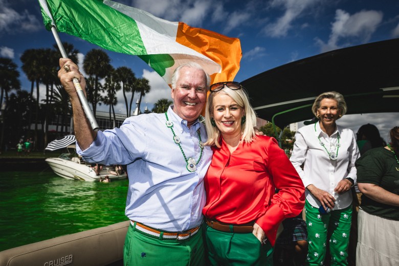 Former Tampa Mayor Bob Buckhorn, an unidentified woman in a red shirt, and current Tampa Mayor Jane Castor posing together at the River-O-Green festival. Buckhorn stands on the left holding an Irish flag, and all three are dressed in festive attire with the bright green dyed Hillsborough River in the background.