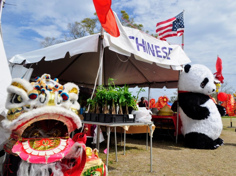 A "Chinese" cultural booth at the 50th Annual SPIFFS International Folk Fair in Pinellas Park. The scene features a large panda mascot, a traditional lion dance head, and a white tent flying an American flag under a bright blue sky.