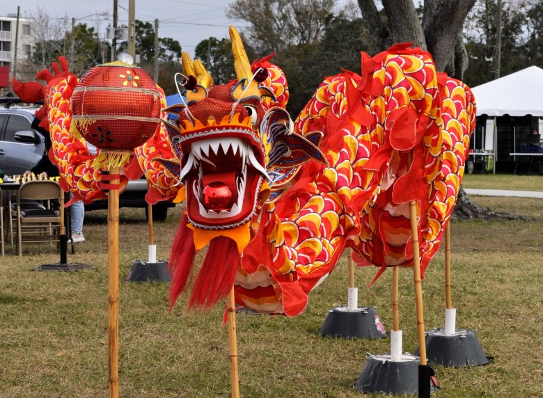 A colorful traditional Chinese dragon puppet on display at a fair hosted by the St. Petersburg International Folk Fair Society (SPIFFS). The large red and yellow dragon features a red pearl in its mouth and is positioned next to a decorative red lantern on a bamboo pole in an outdoor grassy field.