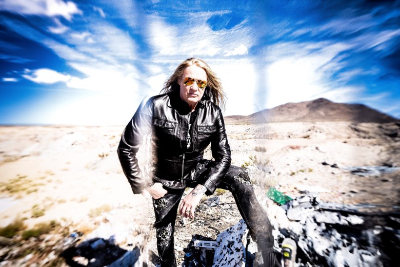 A dramatic wide-angle portrait of metal artist Sebastian Bach in a desert landscape. They are positioned in the center, wearing a black leather jacket, dark distressed jeans with patches, and yellow-tinted aviator sunglasses. They have long blonde hair and a confident expression. The background features a vast, arid desert under a bright blue sky with wispy white clouds, framed by a soft, white vignette and motion-blur effect around the edges.