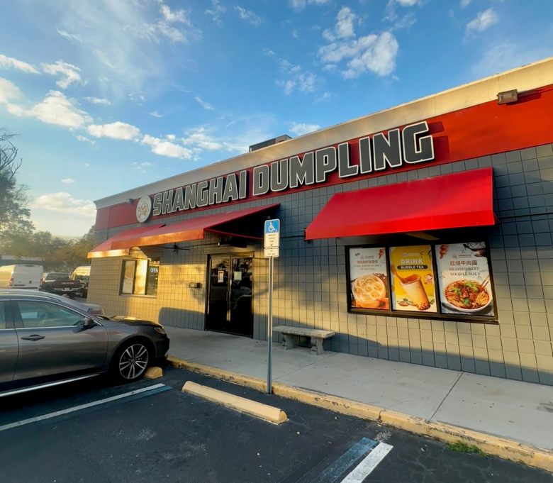 Exterior view of Shanghai Dumpling House on East Fowler Avenue in Tampa. The grey building features a prominent red banner with the restaurant name in white and black block letters, red awnings over the windows and entrance, and large window decals showcasing signature dishes like soup dumplings and noodle bowls under a bright blue afternoon sky.