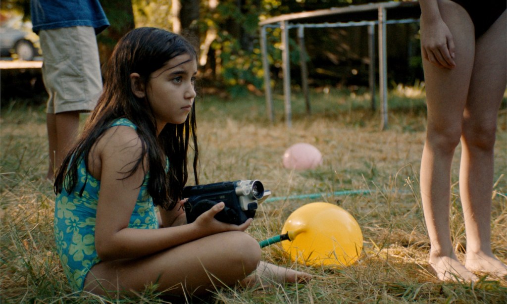 still from the movie 'Blue Heron' showing a young girl in a bathing suit sitting on the grass and holding a camcorder