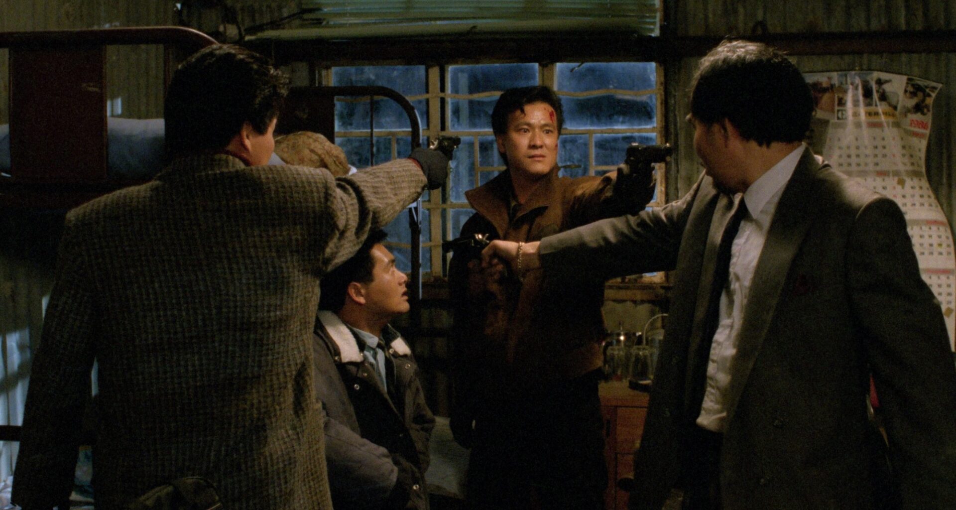 Still from the movie "City of Fire" showing a gun standoff between three men. One of the men points a gun at a man sitting in a chair in the middle of them