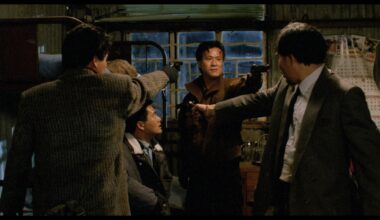Still from the movie "City of Fire" showing a gun standoff between three men. One of the men points a gun at a man sitting in a chair in the middle of them